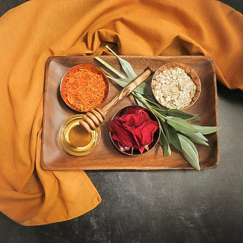 A wooden tray sits on a black surface surrounded by a mustard yellow towel. On the tray there is a copper bowl in the top left corner filled with dry calendula flower petals. Going clockwise on the tray is the handle of a honey wand, fresh sage sprigs, a copper bowl filled with rolled oats, a copper bowl with drie red rose petals and a glass bowl with light colored honey.