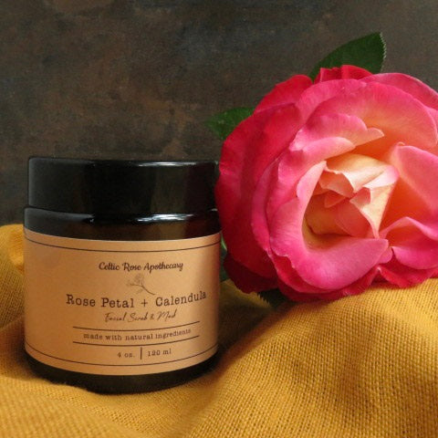 An amber glass jar with Rose petal and calendula face scrub lable sits on a mustard yellow towel next to a pink rose.