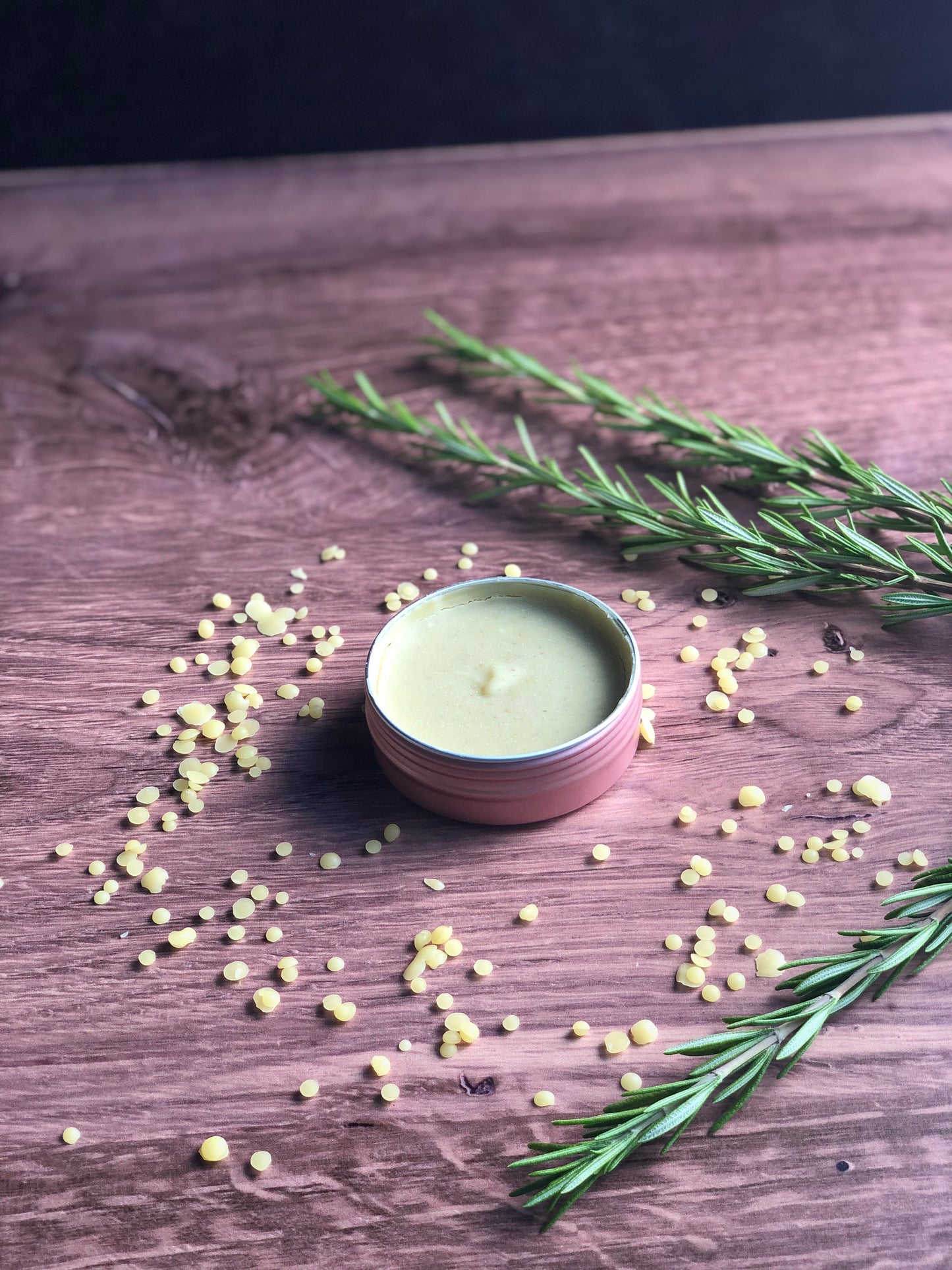 A rose colored salve tin filled with a light green salve sits on a wooden surface. The tin is surrounded by bee's wax pellets and sprigs of fresh rosemary.