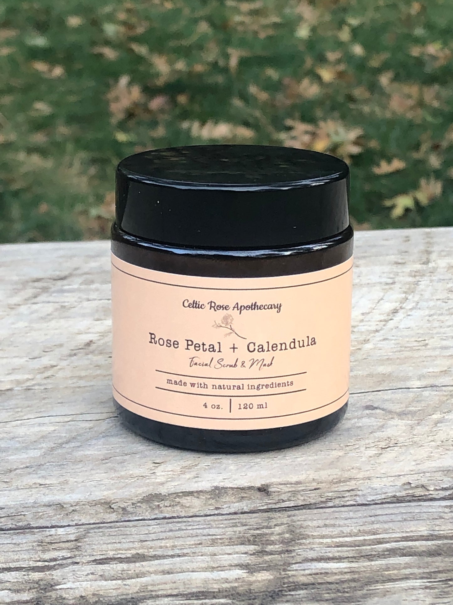 An amber glass jar with a tan lable for rose petal and calendula face scrub sits on a wooden table top. There is green grass in the background.