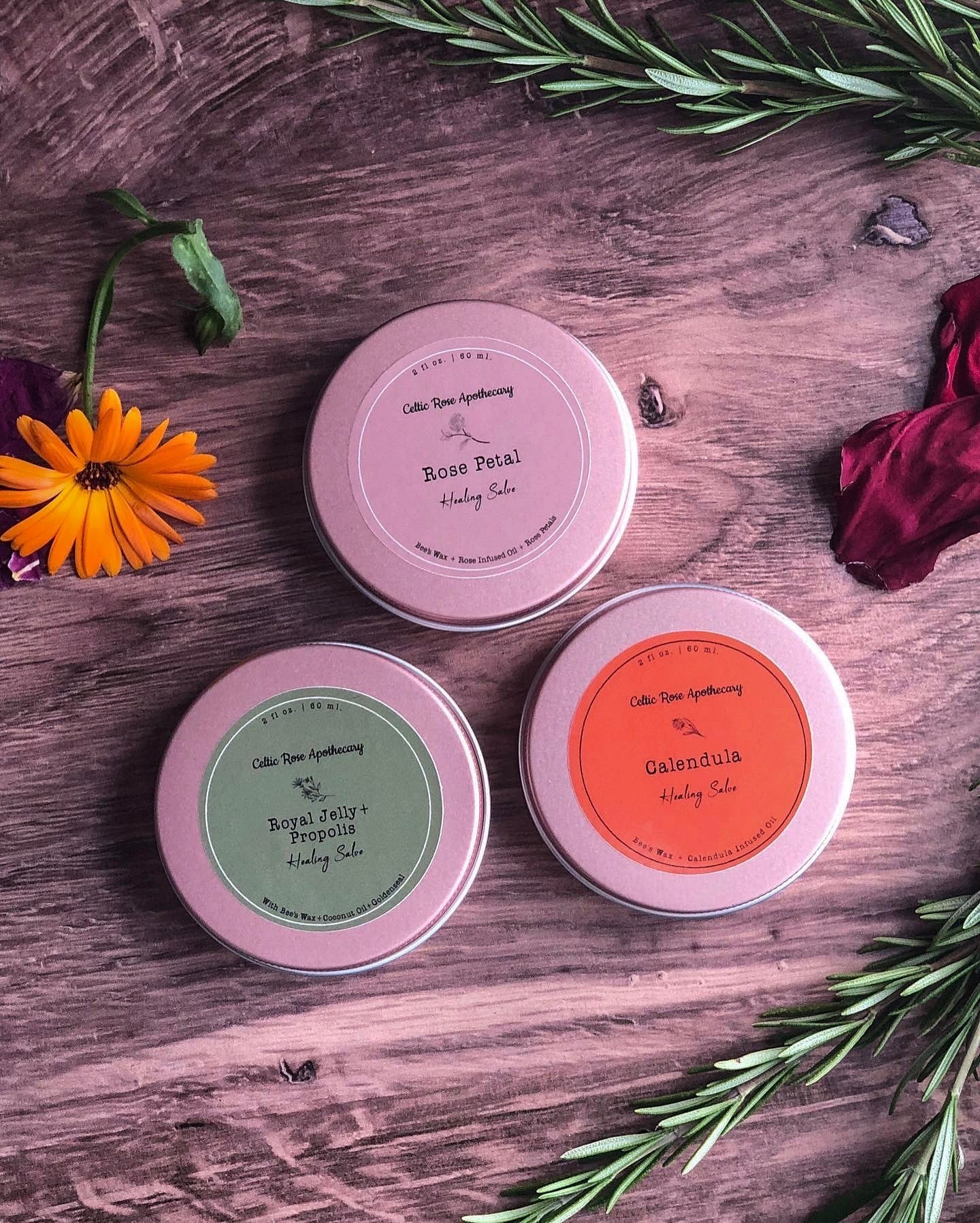 Three rose gold tins sit on a wodden surface surrounded by rosemary clippings, an orange calendula flower, and red dried rose petals. The salves have their coresponding lables attatched to the lid.