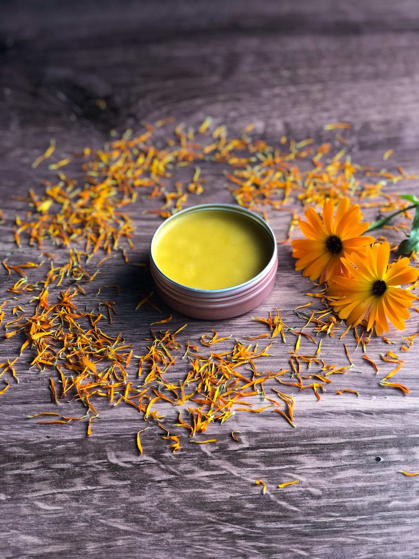 A rose gold coloered tin filled with a light orange salve sits on a wodden surface. The tin is surrounded by orange dried flower petals and two fresh calendula flowers.